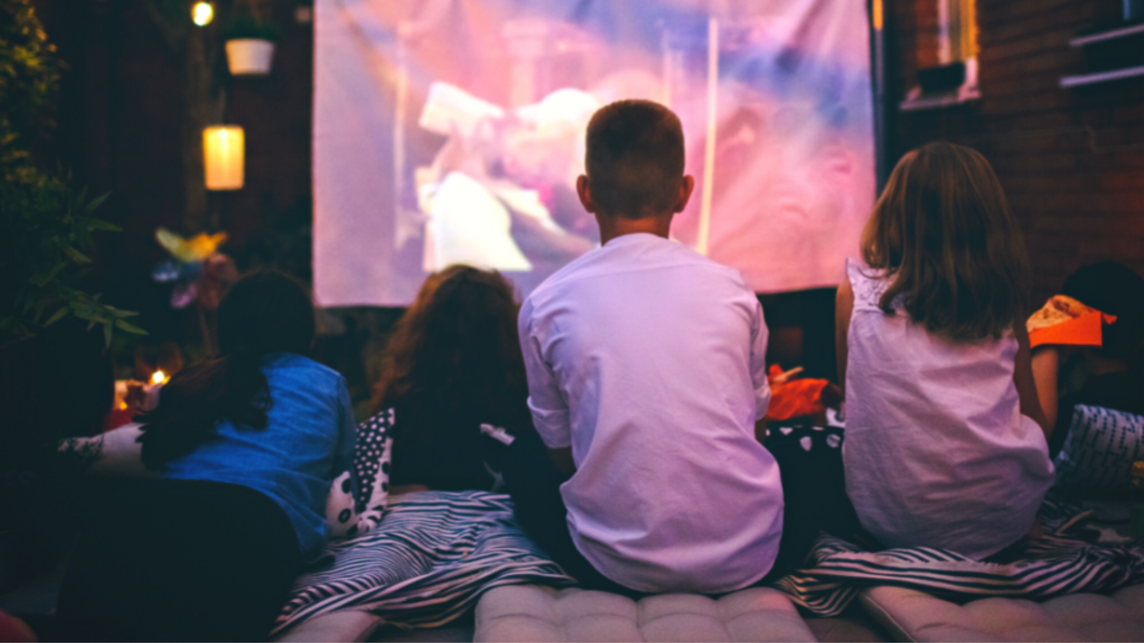 three kids watching a movie on an outdoor screen in the backyard